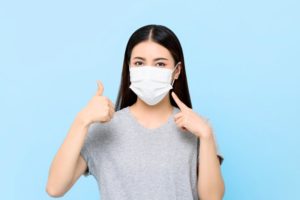 young woman wearing face mask and giving thumbs up against light blue background 