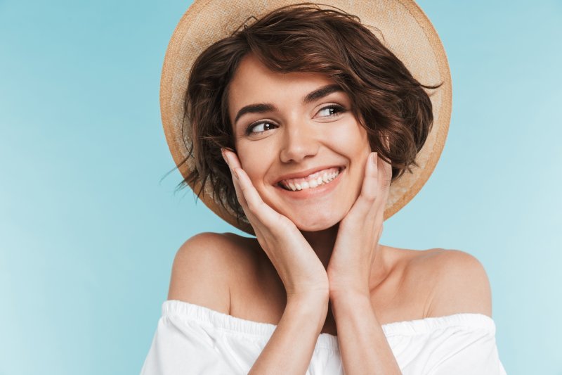 a young woman wearing a hat and holding her face between her hands while smiling and showing off her small teeth in Fanwood